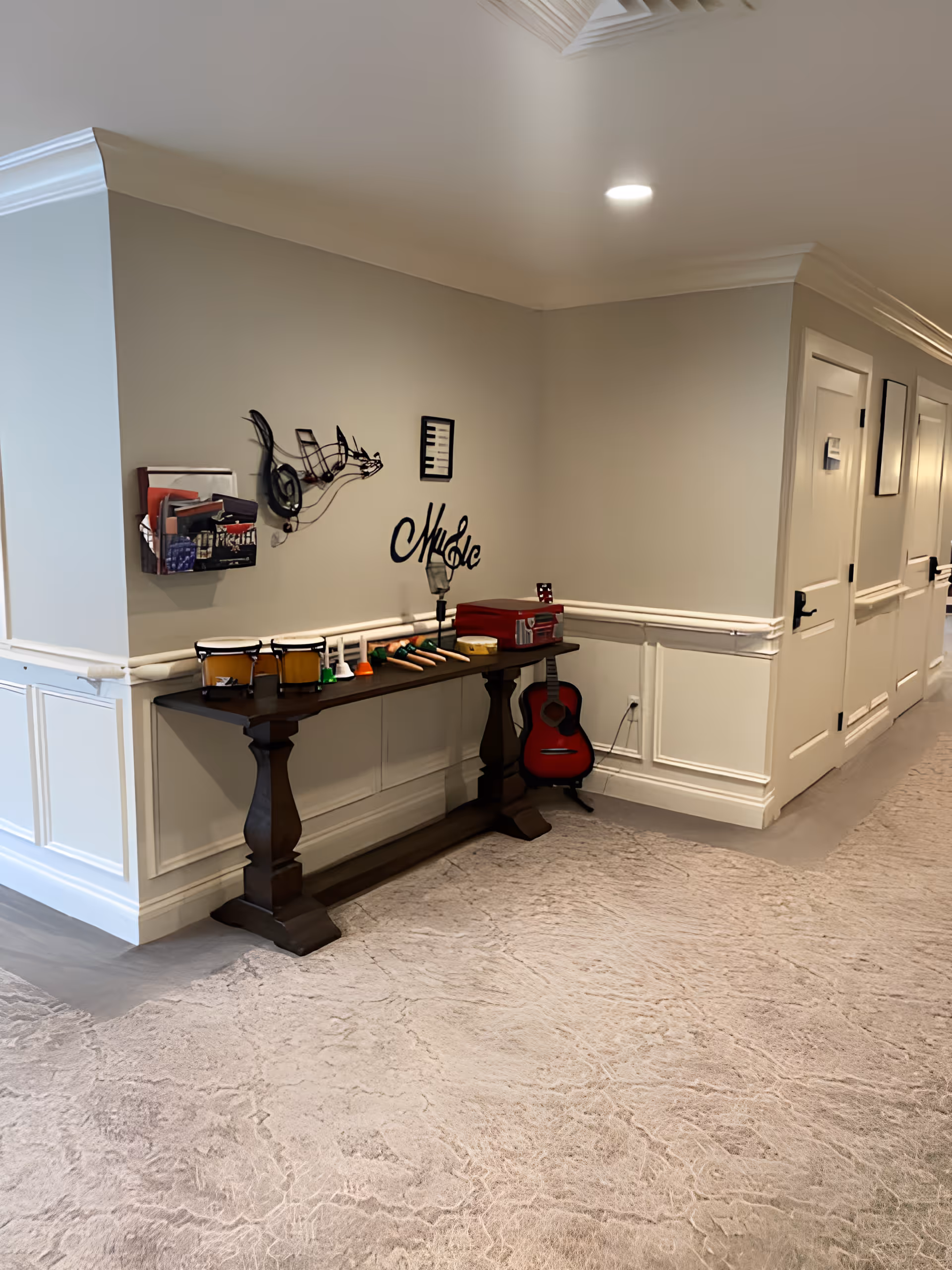 A hallway corner in a senior living facility with a wooden console table against the wall. On the table are various musical instruments including small drums, handbells, and a tambourine. A red acoustic guitar rests on the floor beside the table. The wall above the table features metal wall art shaped like musical notes, a piano keyboard, and the word 'Music'. The hallway has beige walls with white wainscoting and a light-colored carpeted floor.