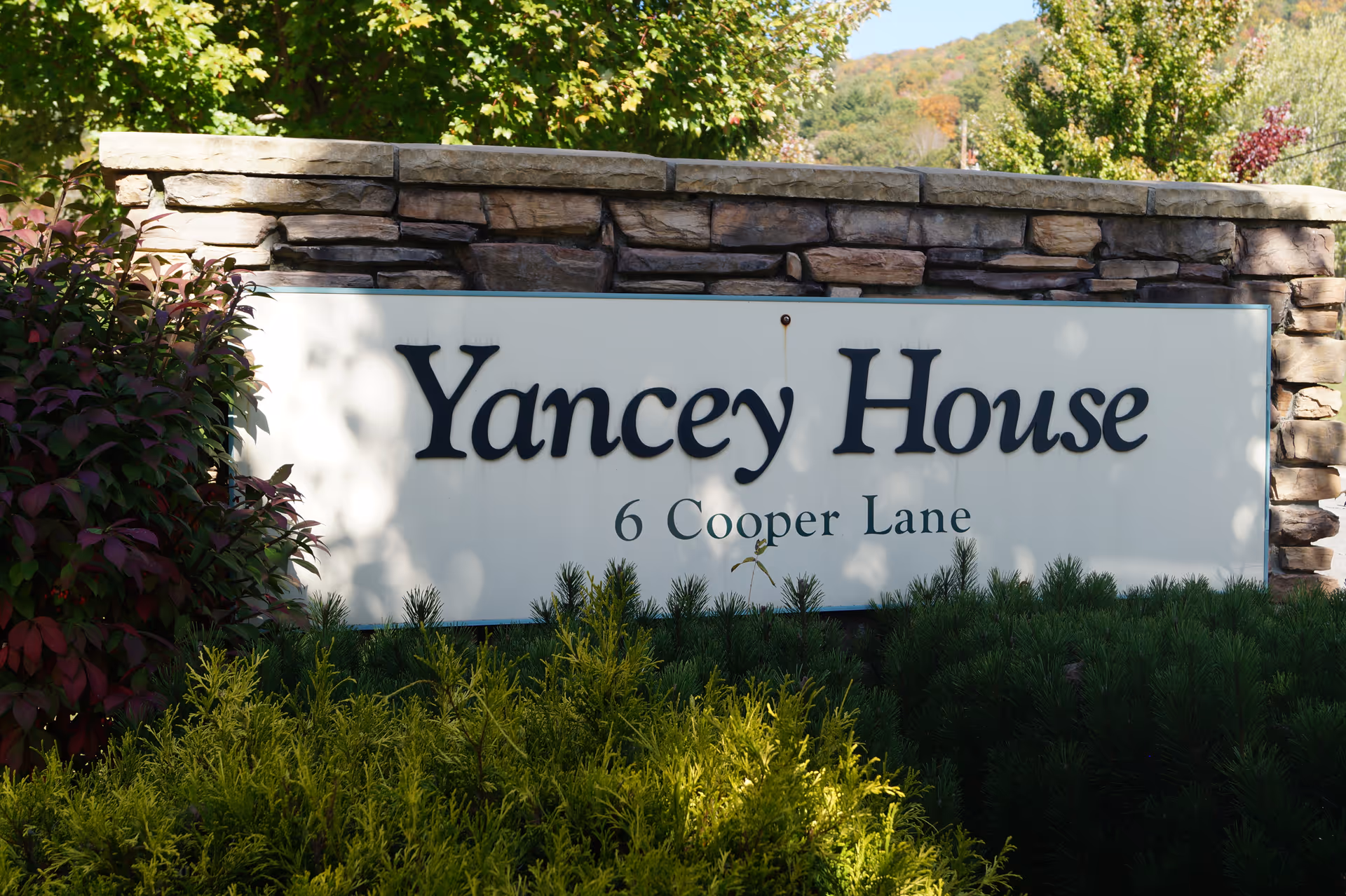 A stone sign with the text 'Yancey House 6 Cooper Lane' surrounded by green bushes and trees with a background of hills and a clear sky.