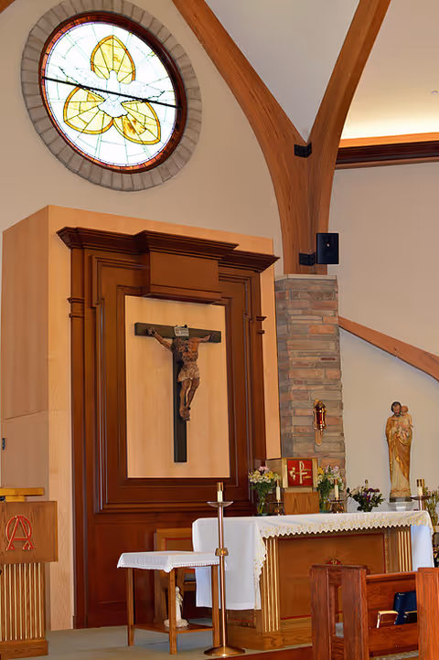 Interior view of a chapel with a wooden altar, a crucifix mounted on a wooden panel, a stained glass window above, and religious statues and flowers around the altar.
