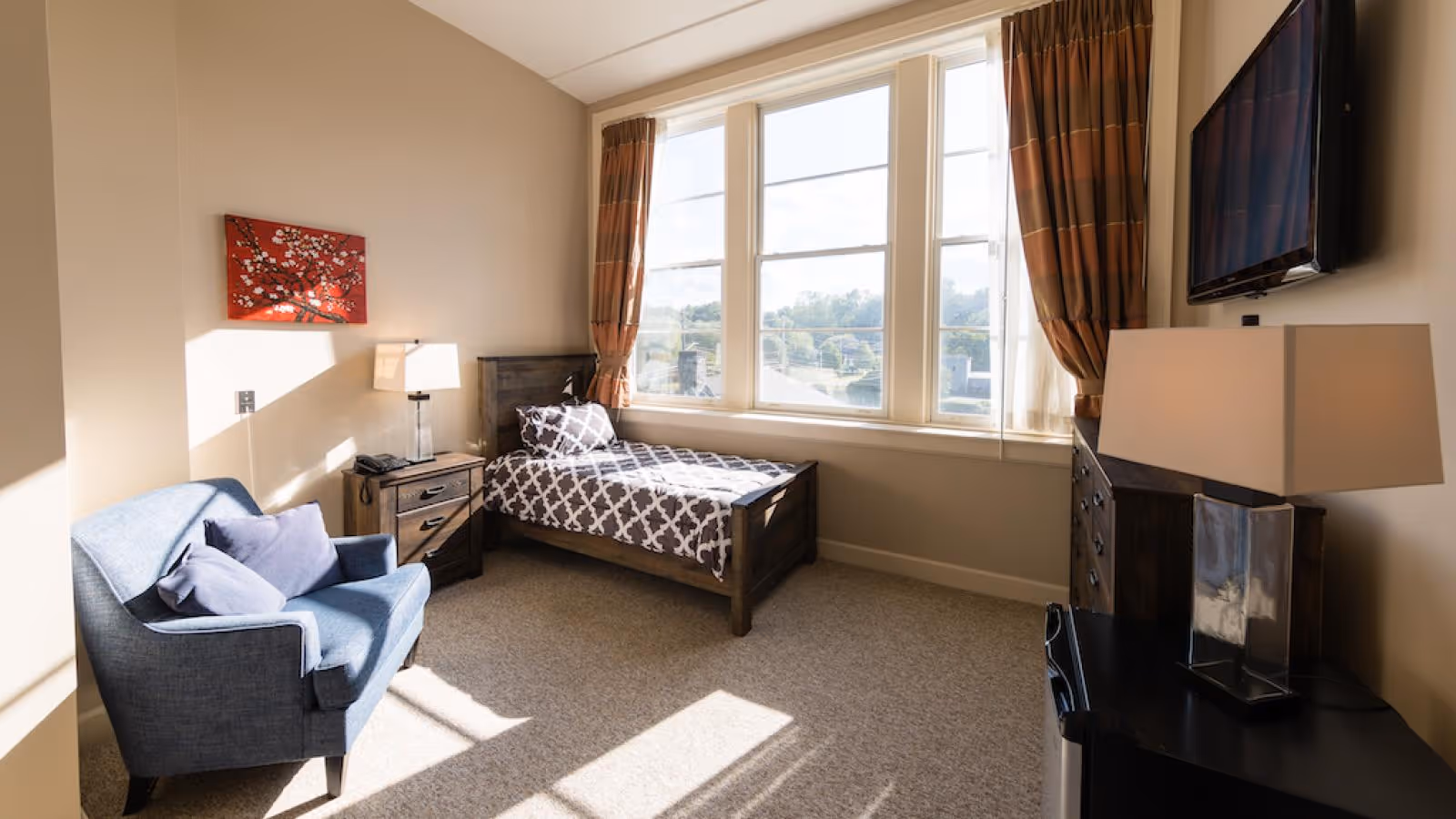 A bright and cozy bedroom in a senior living facility featuring a single bed with patterned bedding, a blue armchair with cushions, a wooden nightstand with a lamp and phone, a dresser with a lamp, and a wall-mounted flat-screen TV. Large windows with brown curtains allow natural light to fill the room.