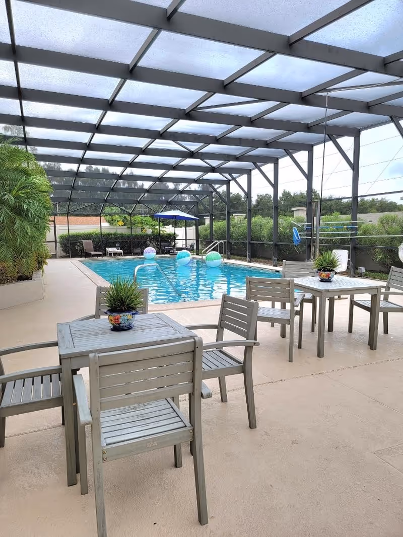 Covered outdoor swimming pool area with several wooden tables and chairs, potted plants on the tables, and colorful beach balls floating in the pool. The area is enclosed with a screened roof and surrounded by greenery.