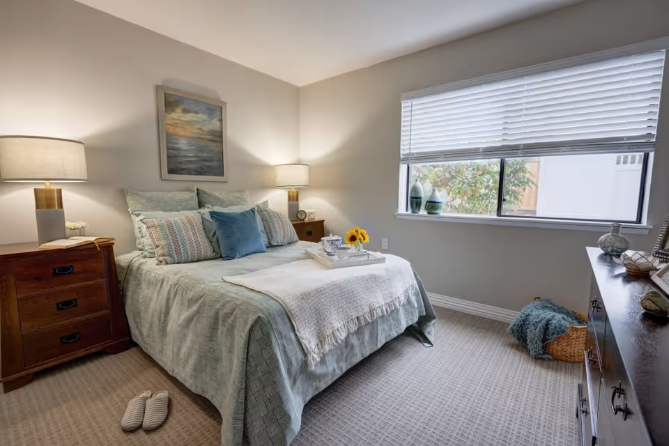 A cozy bedroom with a neatly made bed featuring multiple pillows and a light green bedspread. There are two wooden nightstands on either side of the bed, each with a lamp. A painting hangs above the bed. A window with blinds lets in natural light, and a dresser with decorative items is on the right side. A pair of slippers is placed on the carpeted floor near the bed.