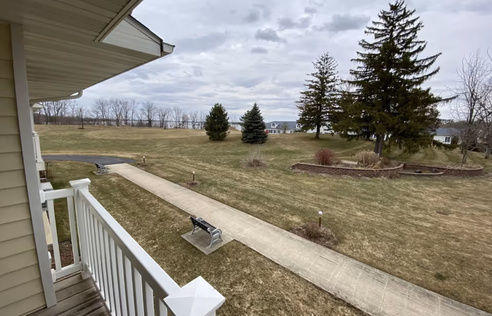 View from a balcony overlooking a grassy outdoor area with a concrete walkway, benches, several trees, and a cloudy sky.