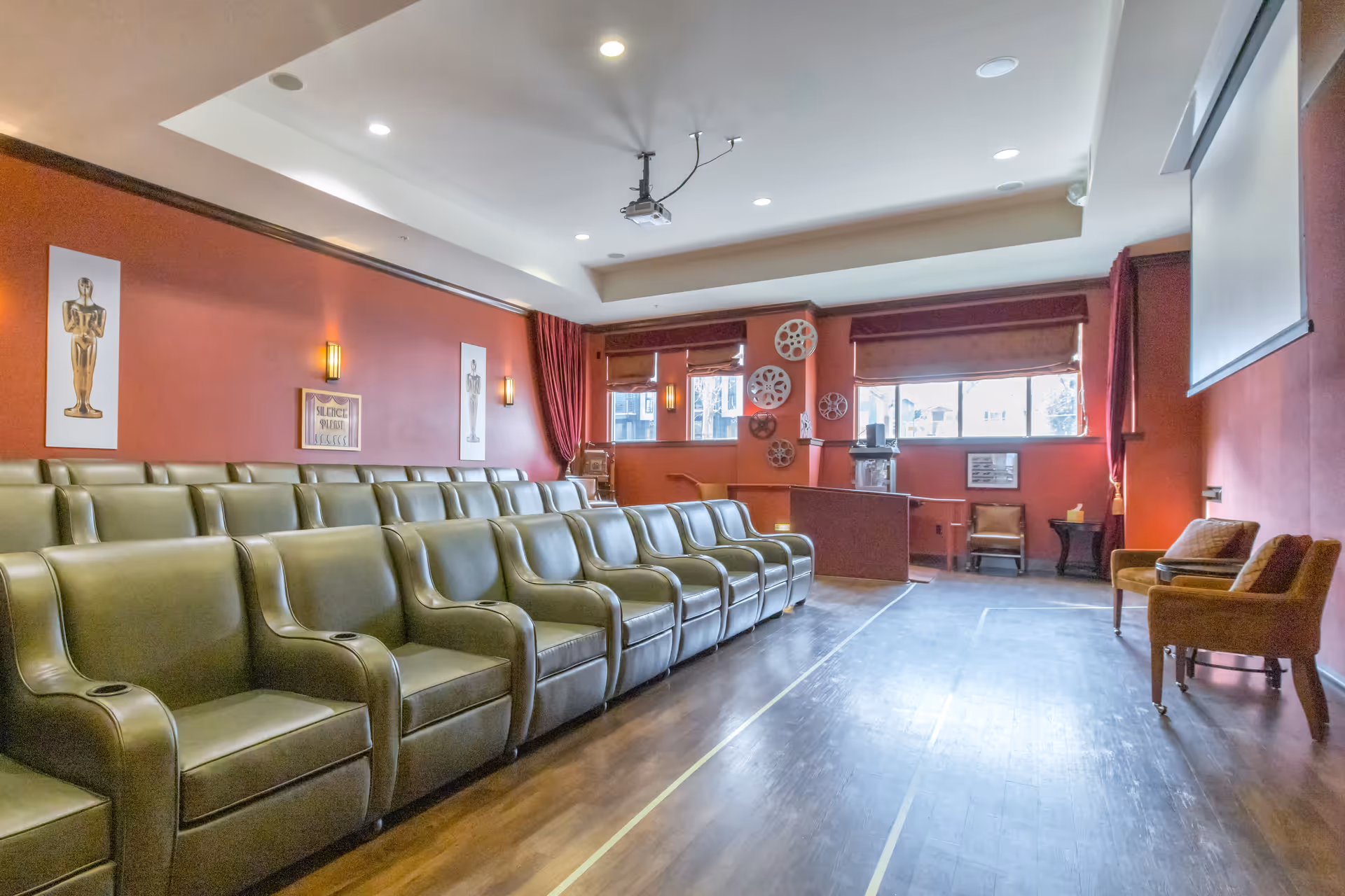 Interior view of a small theater room with rows of green leather armchairs facing a large projection screen. The walls are painted red and decorated with movie-themed artwork and film reels. There are windows with red curtains letting in natural light.
