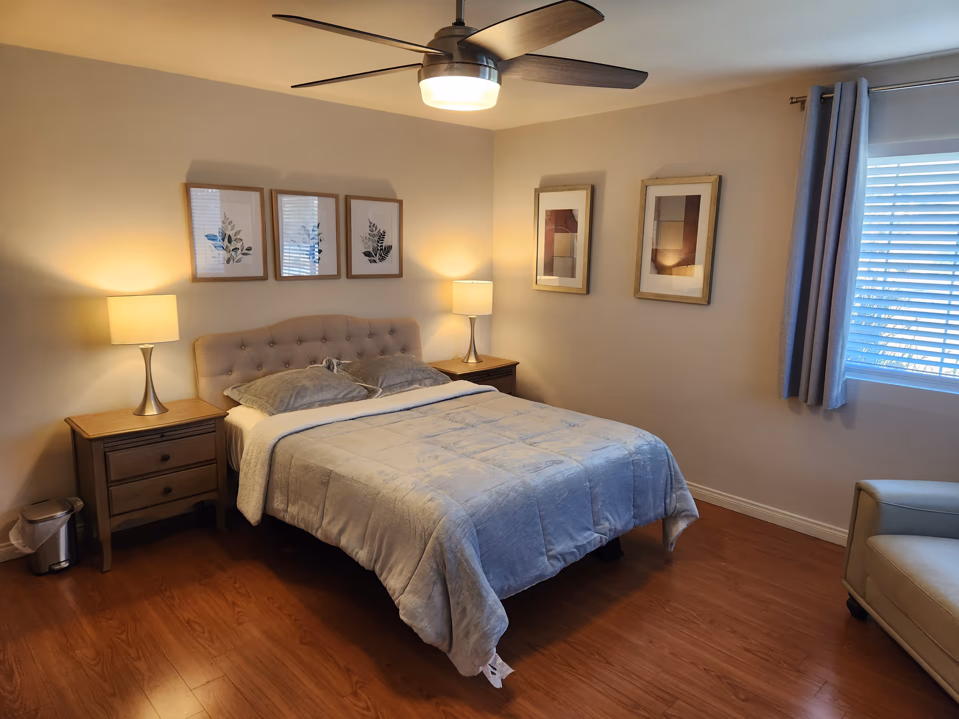 A cozy bedroom with a double bed centered against a beige wall. The bed has a tufted headboard, gray bedding, and two pillows. On each side of the bed are wooden nightstands with matching lamps emitting warm light. Above the bed are three framed botanical prints, and on the adjacent wall are two additional framed abstract artworks. A window with closed blinds and gray curtains is on the right side, and a beige armchair is partially visible in the corner. The room has wooden flooring and a ceiling fan with a light fixture.