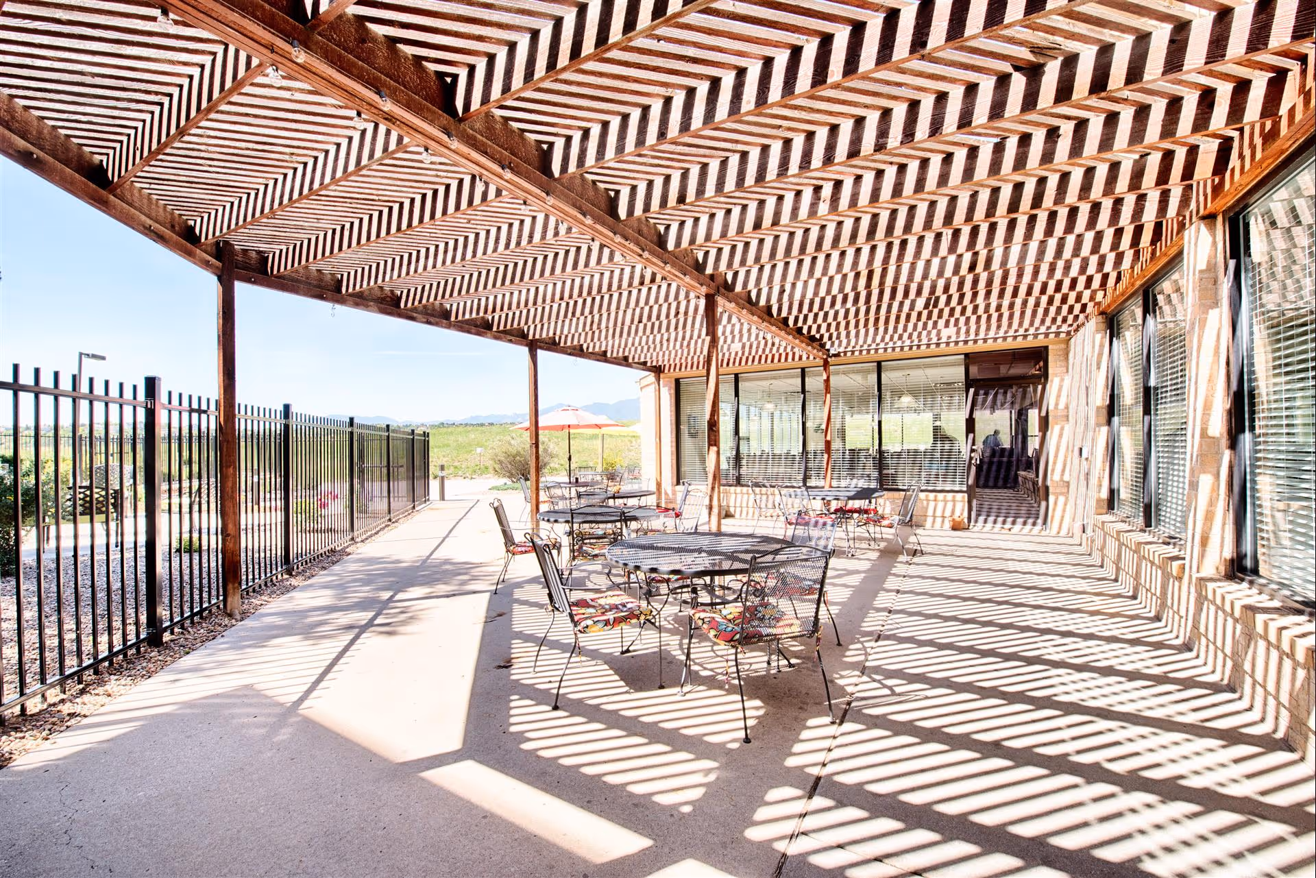 Outdoor patio area with metal tables and chairs featuring colorful cushions, shaded by a wooden pergola casting striped shadows on the concrete floor. A black metal fence runs along one side, and large windows with blinds are on the building side.
