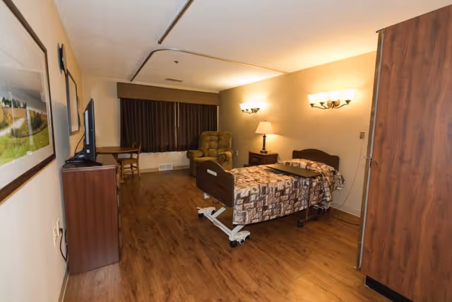 A senior care facility bedroom with a hospital-style bed covered in a patterned blanket, a reclining chair near the window with closed dark curtains, a wooden nightstand with a lamp, a small table with a chair, a flat-screen TV on a wooden cabinet, and wood flooring.