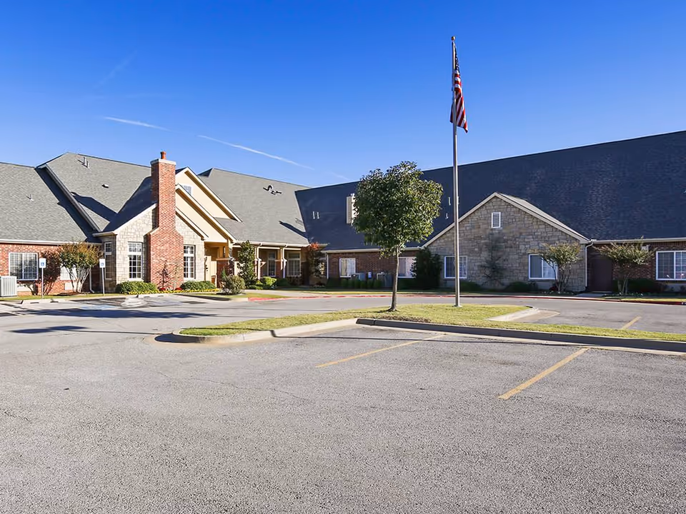 Exterior view of a single-story assisted living facility building with a large parking lot in front, a flagpole with an American flag, and a clear blue sky.