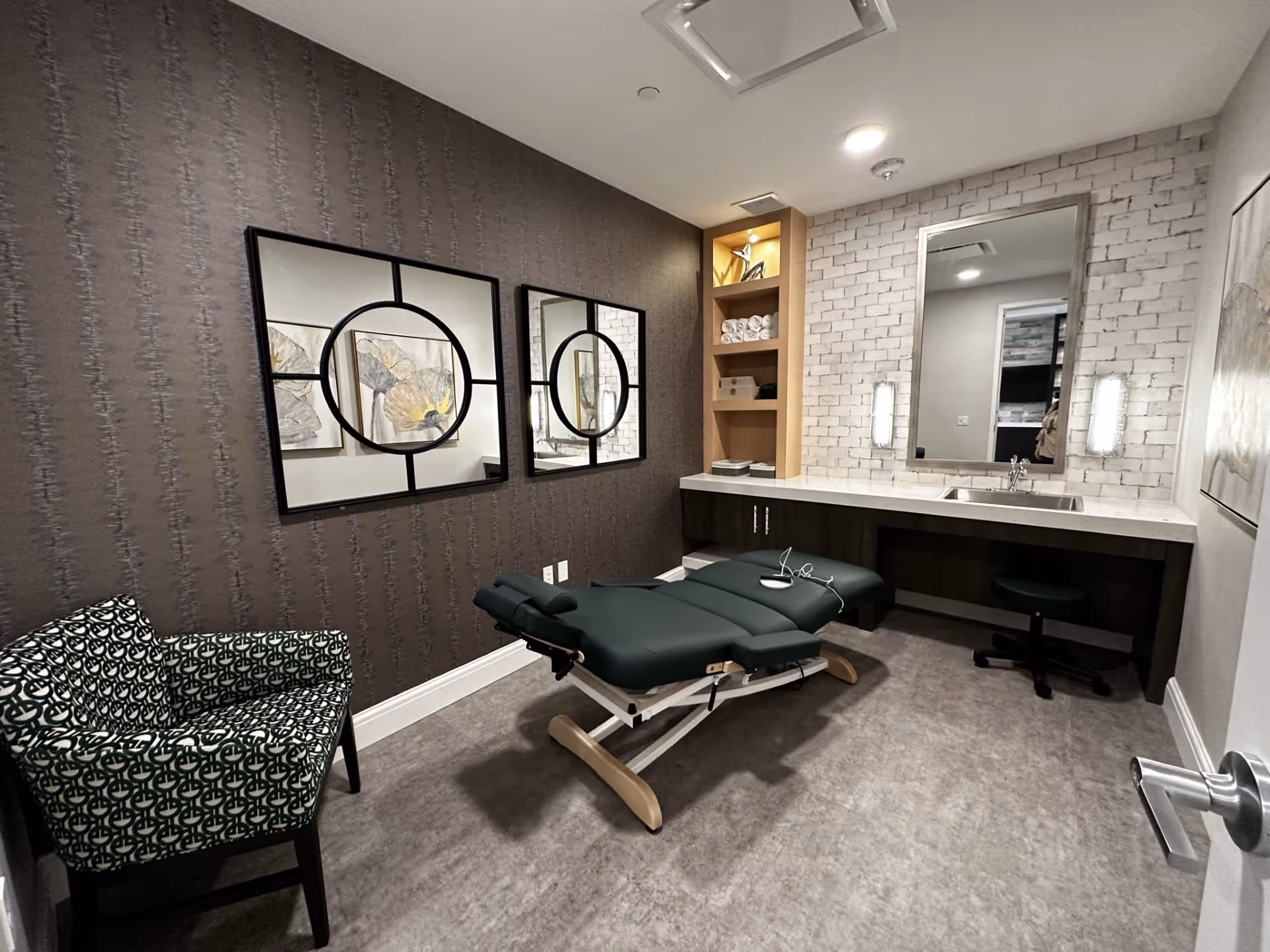 A treatment room with a padded adjustable massage or therapy table in the center, a patterned armchair to the left, two decorative mirrors on a dark textured wall, and a white brick accent wall with a large mirror above a countertop with a sink and shelves holding towels and decor items.