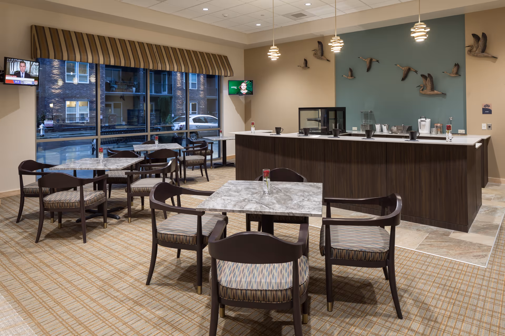 A dining area with several square marble-top tables and wooden chairs with patterned cushions. In the background, there is a counter with coffee and beverage supplies, and a wall decorated with mounted bird sculptures. Two small televisions are mounted on the walls near large windows with a striped valance, showing an outside view of a building and parked cars.