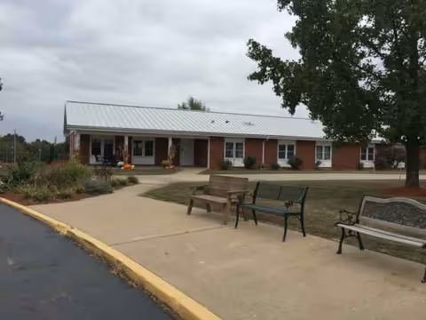 Single-story brick senior living building with a wide entrance, walkway and benches outside.