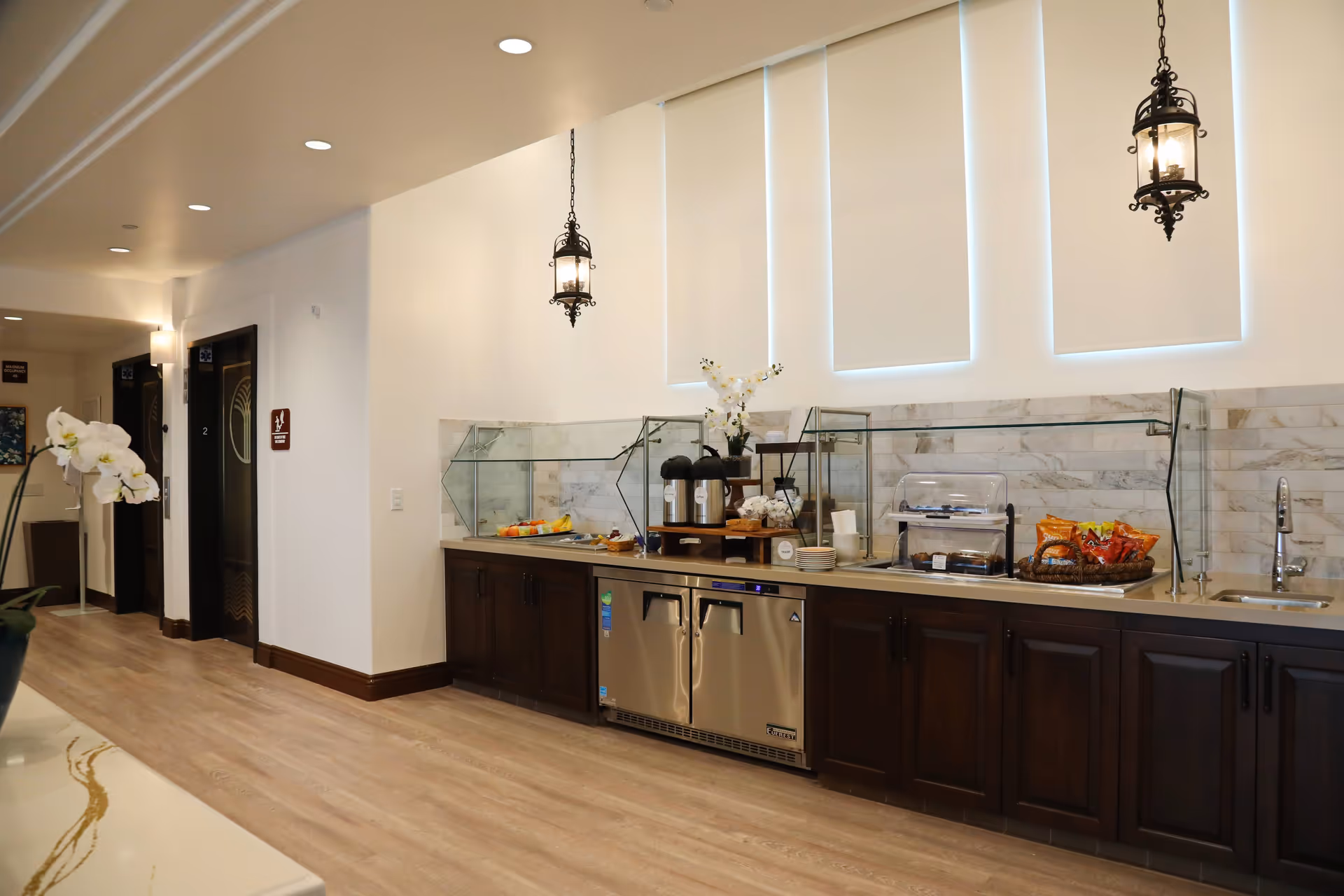 A well-lit interior area with a long counter featuring a coffee station, snacks, and a small sink. The counter has dark wood cabinets and a marble backsplash. Two decorative hanging lantern-style lights are above the counter. In the background, there are two elevators with dark doors and a hallway with light wood flooring and white walls.