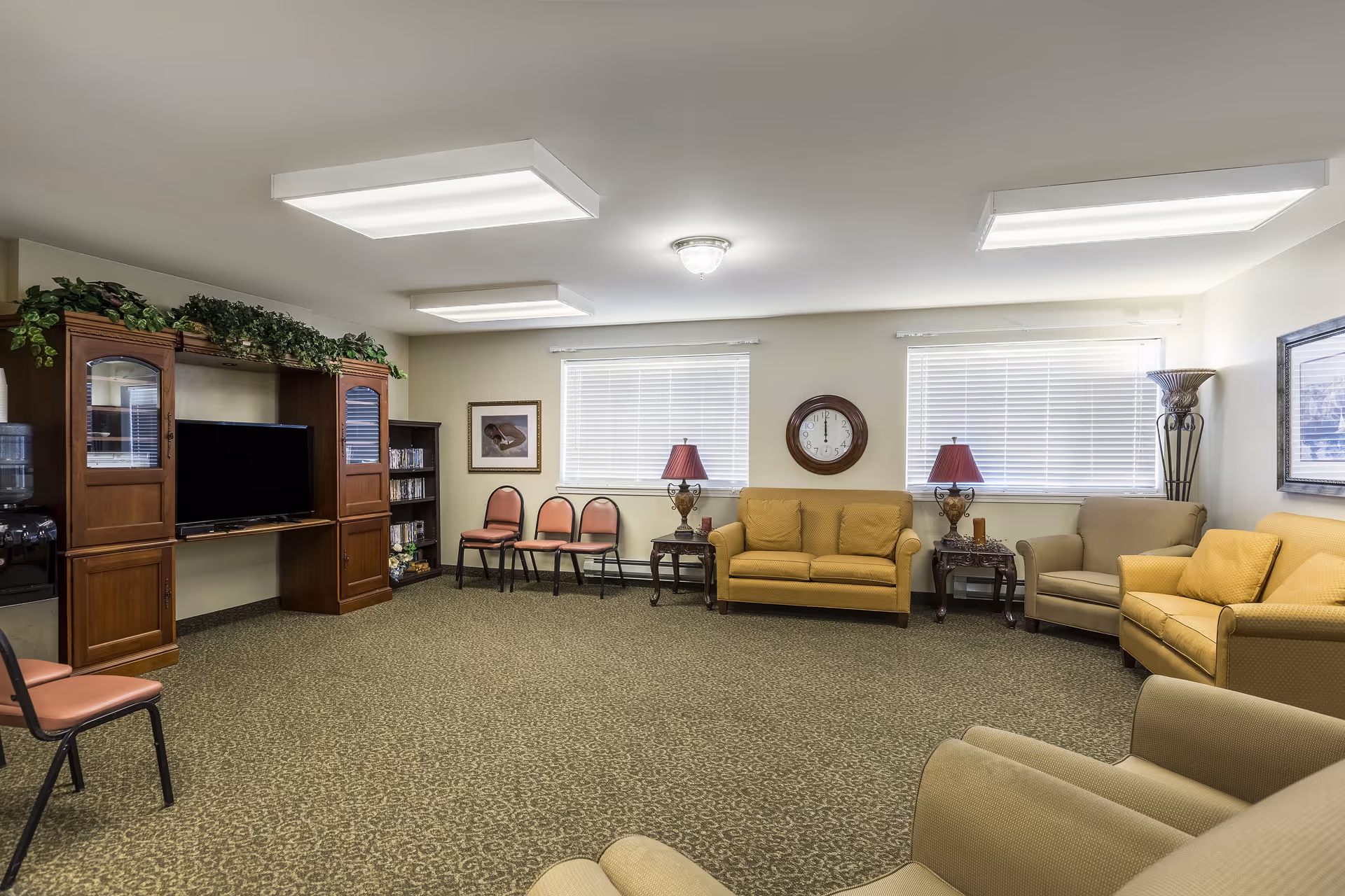 A spacious senior living community lounge with beige and yellow upholstered armchairs and sofas arranged around the room. There are two windows with white blinds, a wall clock, two table lamps with red shades on wooden side tables, and a wooden entertainment center with a TV and shelves filled with DVDs. The carpet is patterned in green tones, and the ceiling has bright rectangular fluorescent lights.