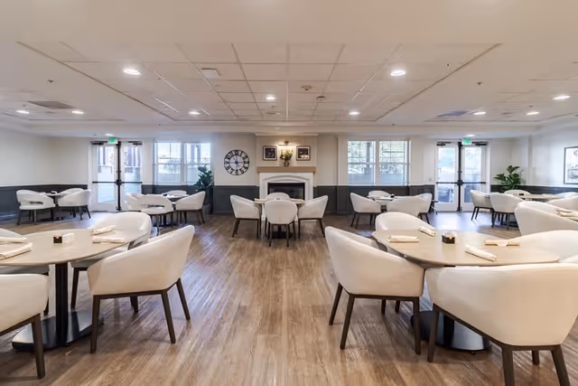 A spacious dining room with multiple round tables surrounded by white cushioned chairs. The room features wood flooring, a fireplace centered on the far wall with framed pictures above it, large windows letting in natural light, and double glass doors on either side. The ceiling has recessed lighting and the walls are painted white with a dark wainscoting.