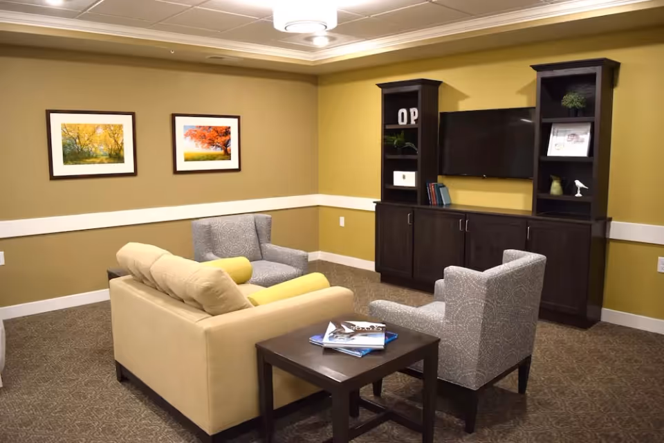 A cozy living room with a beige couch, two patterned gray armchairs, and a dark wooden coffee table with magazines on it. The walls are painted yellow with white trim, and two framed pictures of autumn trees hang on the wall. A dark wooden entertainment center with shelves and cabinets holds a flat-screen TV and decorative items.