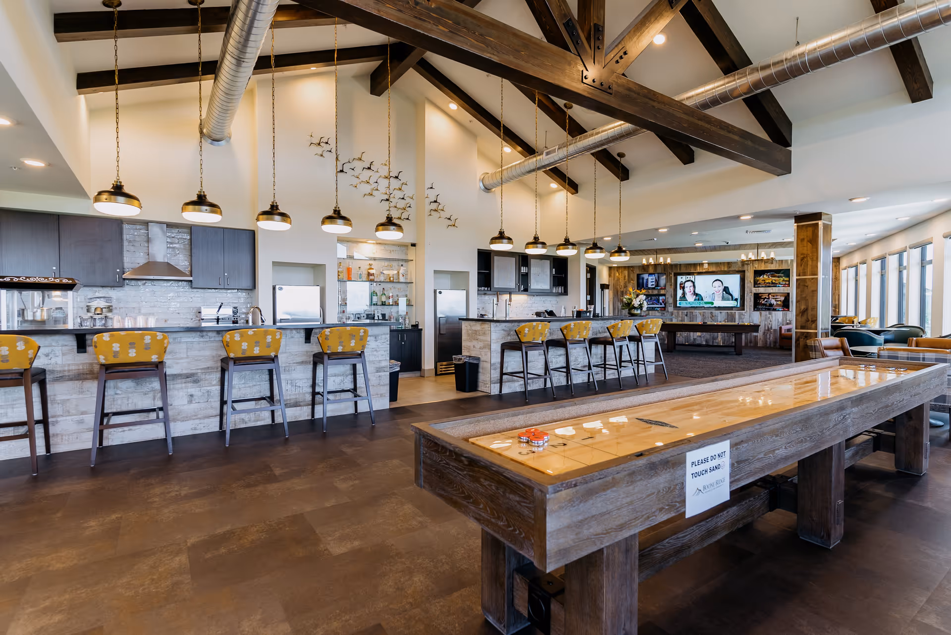 A spacious and modern common area in Boone Ridge Retirement Community featuring a long bar counter with yellow cushioned bar stools, pendant lights hanging from a high ceiling with exposed wooden beams and ductwork, a shuffleboard table with a sign that reads 'Please do not touch sand', and a lounge area with a large TV and comfortable seating near large windows.