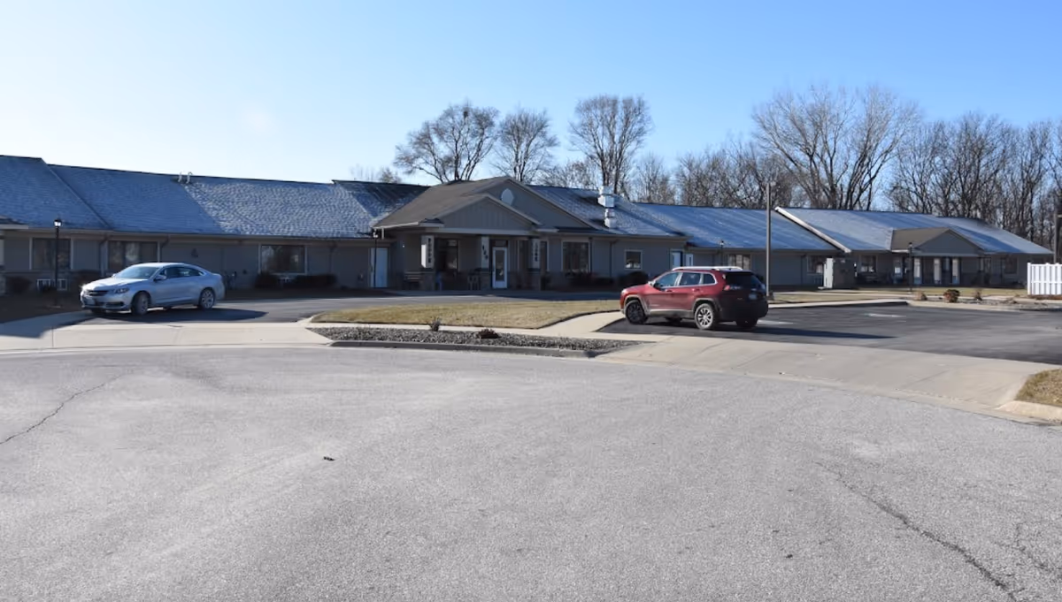 Single-story assisted living building with a central entrance, parking area and two cars under a clear blue sky.