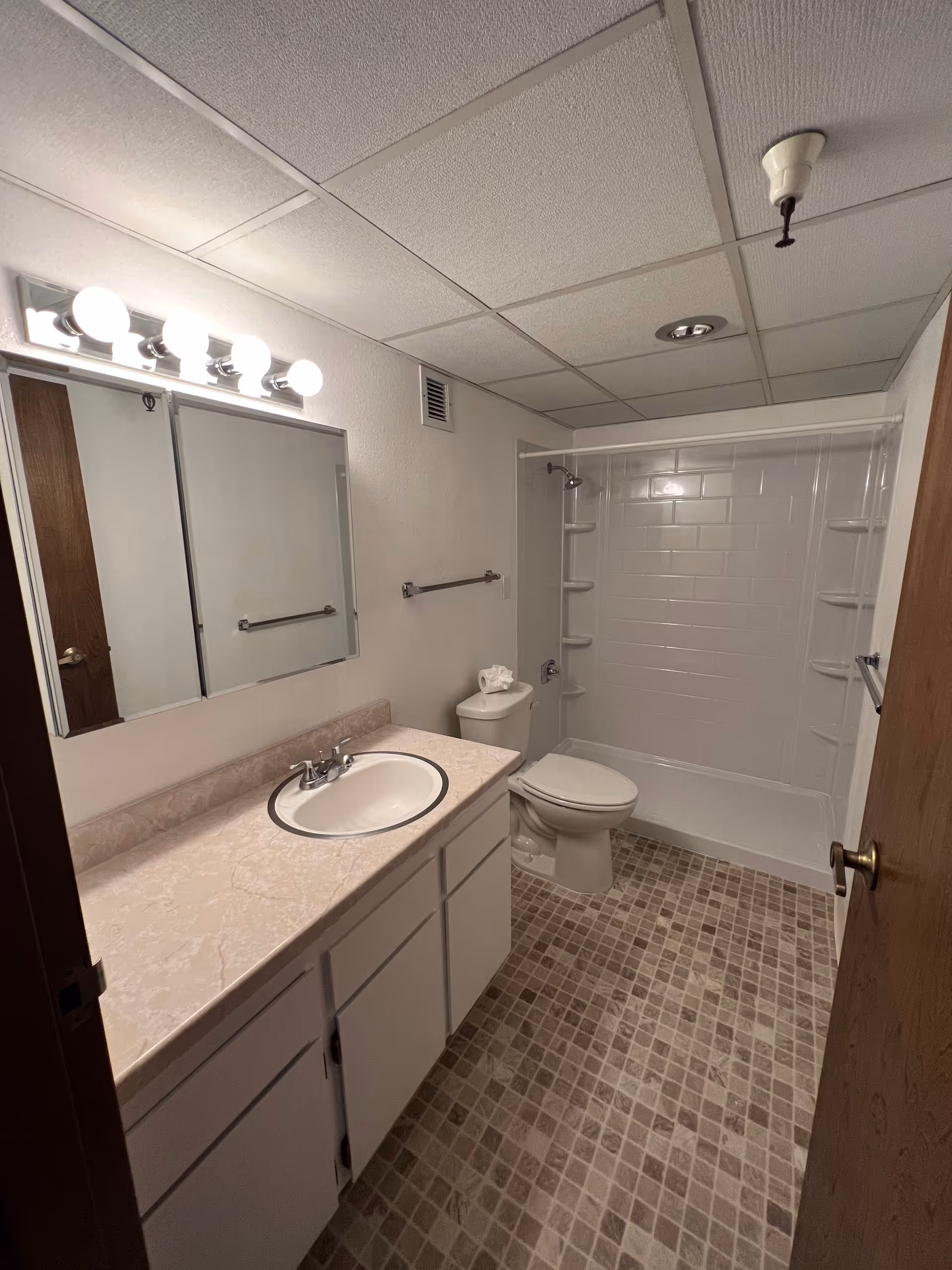 A clean bathroom with a white sink and countertop, a large mirror with four round lights above it, a toilet, and a shower area with built-in shelves. The floor has a tiled pattern in neutral tones, and the walls are painted white.