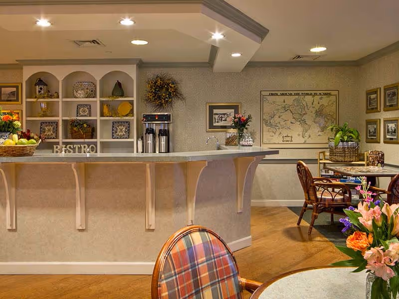 Interior view of a cozy bistro area in a senior living facility featuring a counter with coffee dispensers, decorative shelves with plates and ornaments, a table with a floral centerpiece, and framed artwork on the walls.