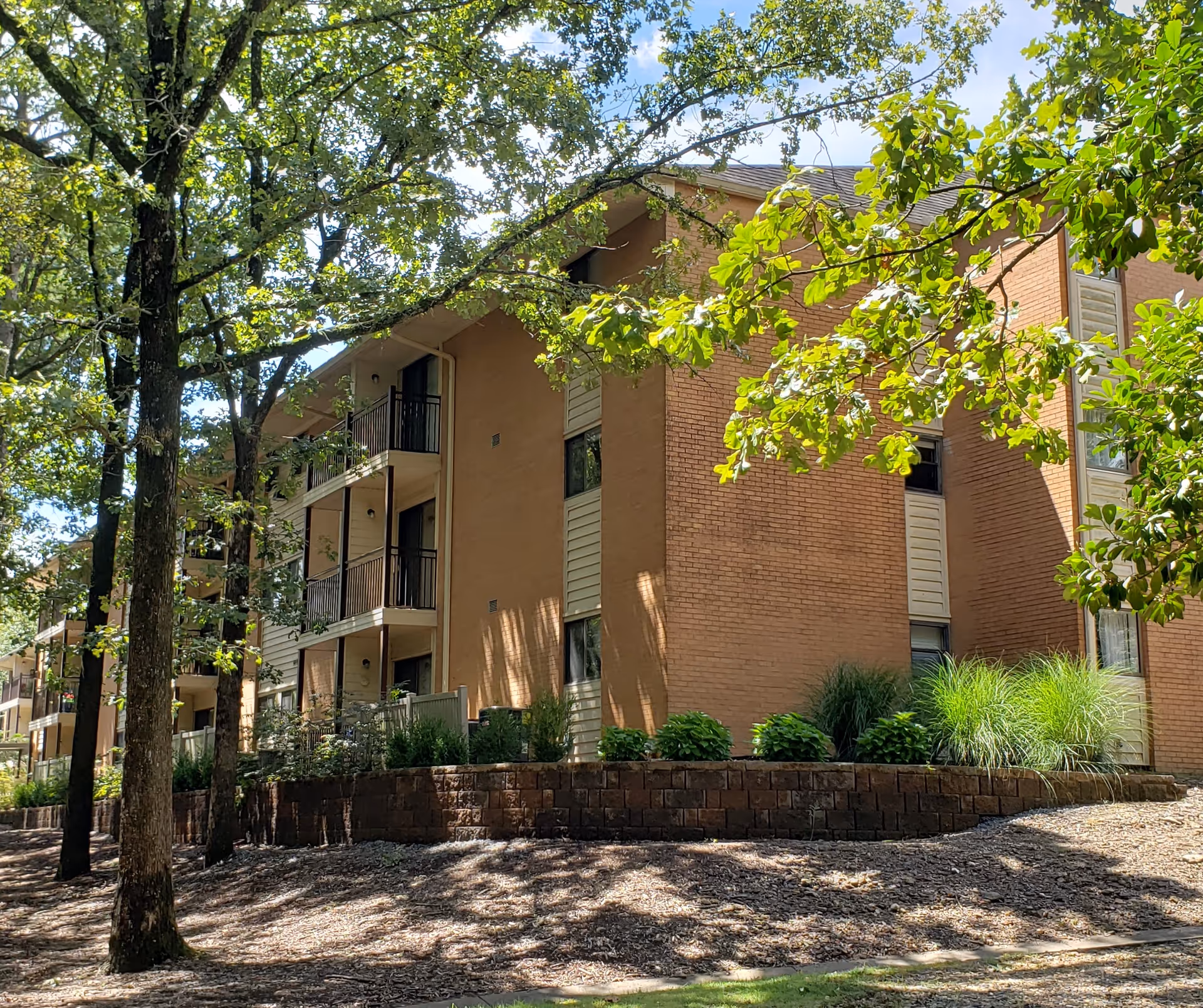 Exterior view of a multi-story brick building with balconies, surrounded by trees and landscaping under a sunny sky.