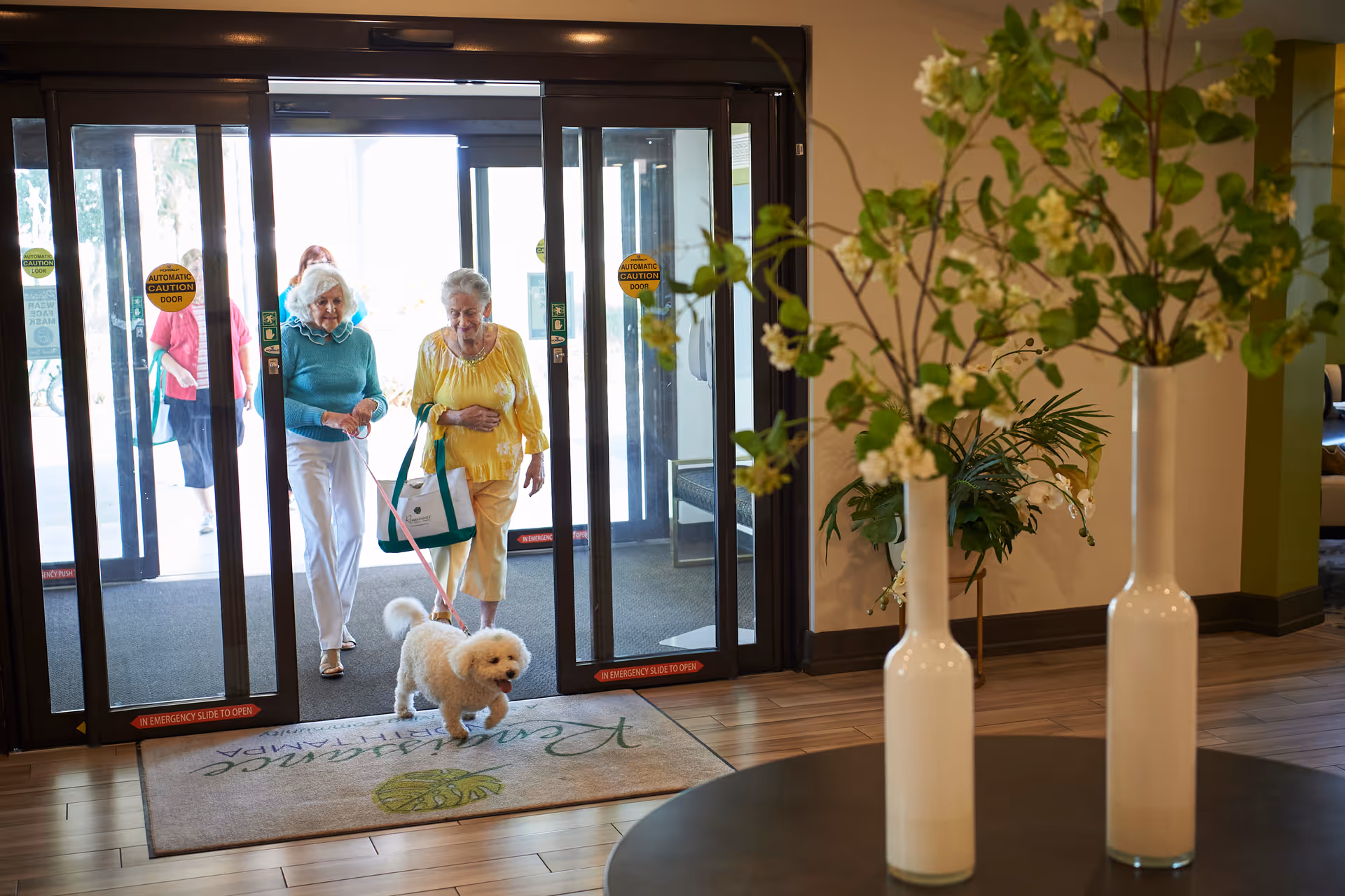 Two elderly women entering through automatic glass doors into a facility lobby, one holding a small white dog on a leash. The floor has a welcome mat with the text 'Renaissance North Tampa'. In the foreground, there are two white vases with green leafy plants on a round table.