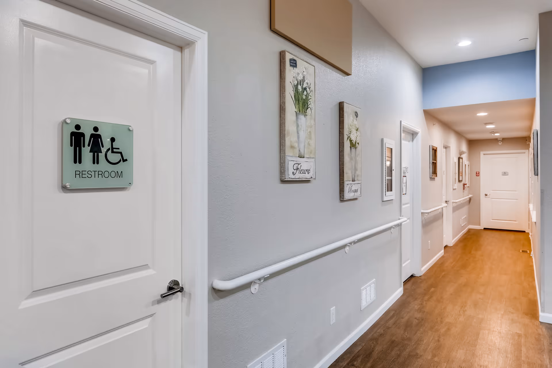 A clean, well-lit hallway with light gray walls and wood flooring. On the left side, there is a white door with a restroom sign indicating it is accessible for men, women, and people with disabilities. The hallway has handrails along the walls and decorative floral artwork hanging above the handrails.