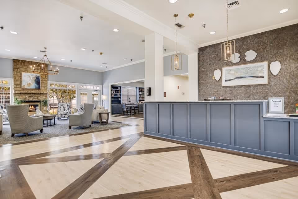A spacious senior living facility common area featuring a reception desk with pendant lights, a seating area with armchairs around a coffee table, a stone fireplace, large windows letting in natural light, and a patterned wooden floor.