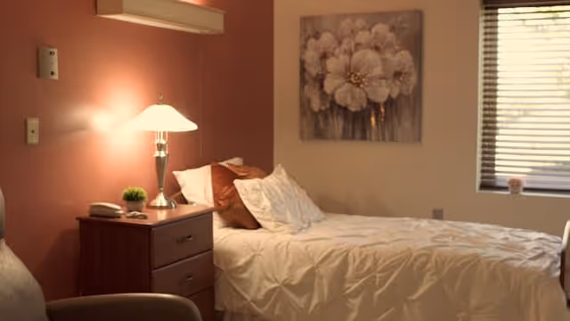 A cozy assisted living bedroom with a made bed, bedside table and lamp, floral wall art, and a window with blinds.