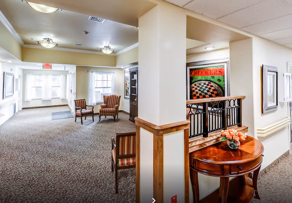 Well-lit communal hallway seating area with armchairs, a side table with flowers, and framed artwork inside a memory care facility.