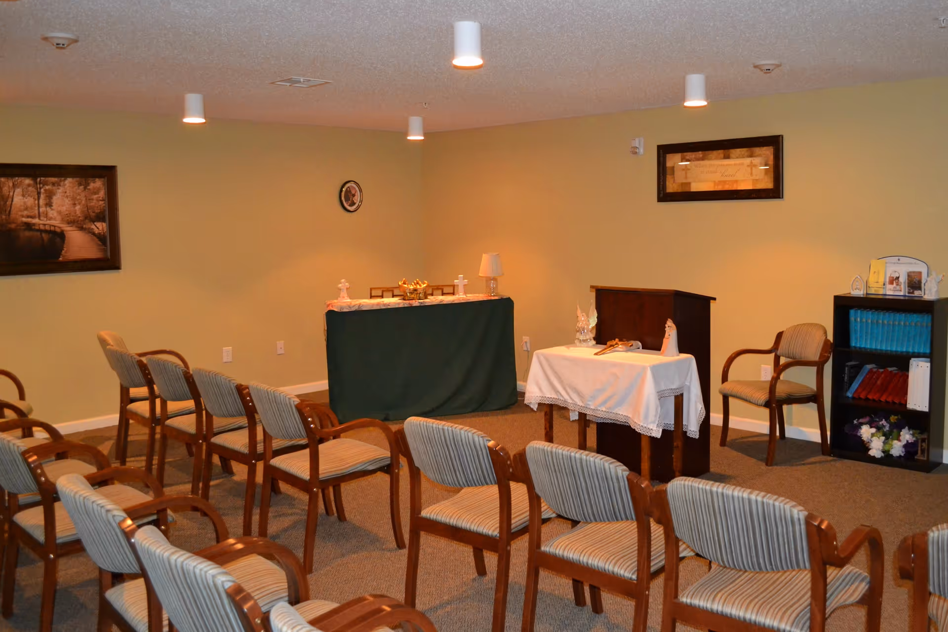 A small chapel or meditation room with rows of wooden chairs with striped cushions facing two tables covered with cloths, one green and one white, adorned with religious items including crosses and candles. The room has beige walls with framed pictures and a small bookshelf with books and flowers.