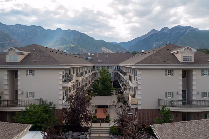 View of a senior living facility named Escalante at Coventry with multiple beige buildings surrounding a central courtyard with trees and a covered seating area, set against a backdrop of green mountains under a cloudy sky.