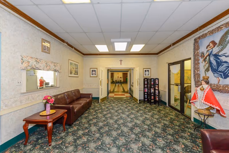 Carpeted lobby/lounge with leather sofas, a side table with flowers, religious artwork, and double doors opening to a hallway.