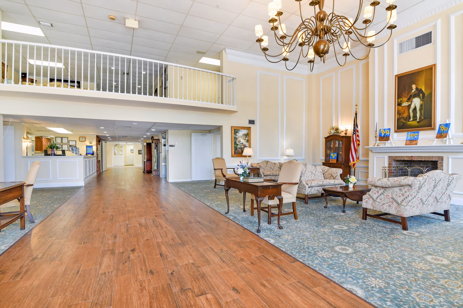 Spacious and well-lit assisted living facility common area with wooden flooring and a large patterned carpet. The room features a seating arrangement with floral upholstered sofas and armchairs around wooden coffee tables. A large chandelier hangs from the ceiling, and there is a fireplace with a portrait above it. The walls are decorated with framed artwork and an American flag stands near a wooden cabinet. A reception desk is visible in the background along with a hallway leading further into the building.