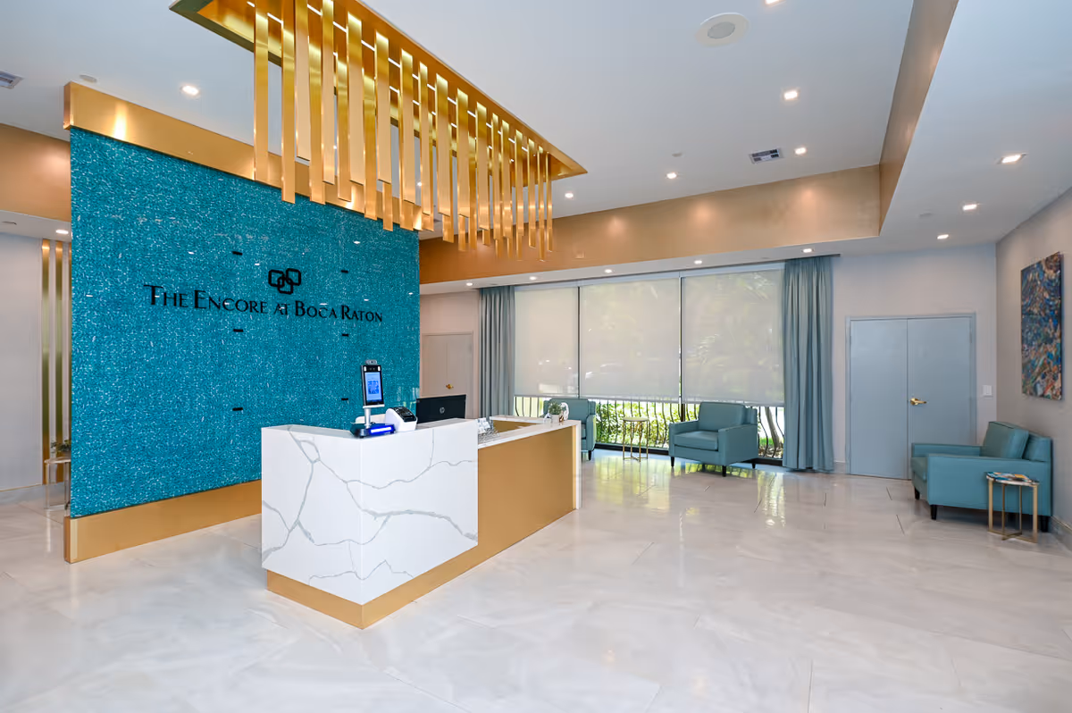 Bright reception lobby with a white marble front desk, turquoise accent wall reading 'The Encore at Boca Raton', gold ceiling fixtures, and teal seating.