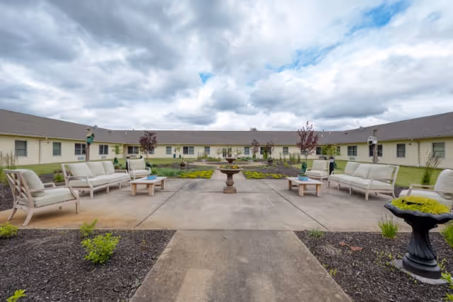 Outdoor courtyard area at Cedarhurst Senior Living of Granite City featuring cushioned patio seating arranged around a central concrete walkway with a decorative fountain in the middle. The courtyard is surrounded by a single-story building with multiple windows and a cloudy sky overhead.