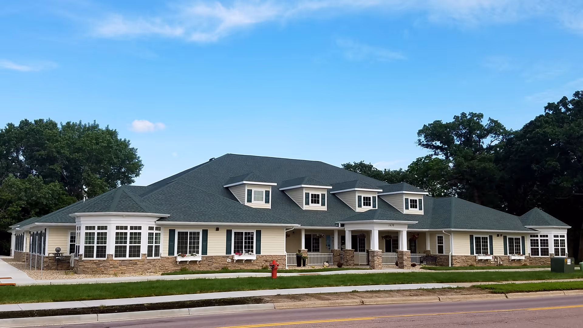 Large single-story cream-colored senior living building with a green roof, multiple dormer windows, a covered entrance porch, and a lawn facing the street.