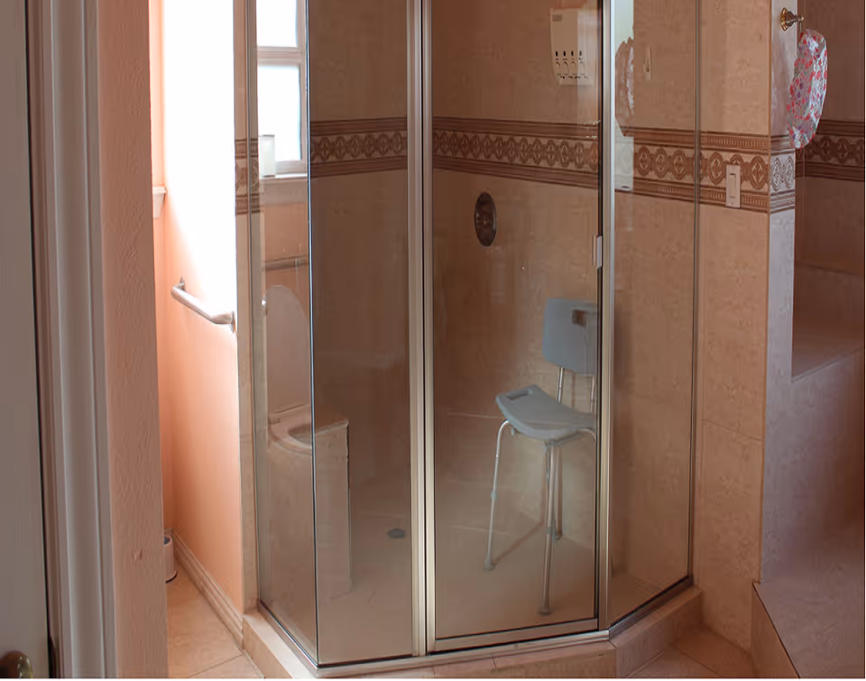 A bathroom featuring a glass-enclosed shower with a gray shower chair inside. The walls and floor are tiled in beige tones with a decorative border tile. There is a window with frosted glass and a grab bar mounted on the wall near the toilet, which is partially visible.