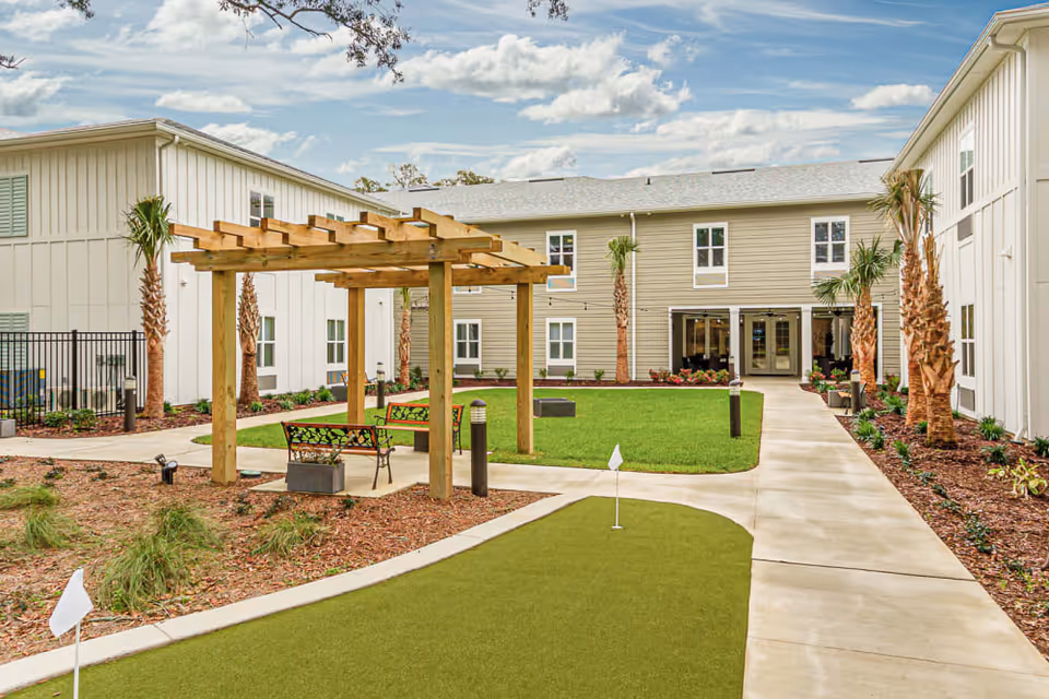 Outdoor courtyard area at Arcadia Senior Living Pace featuring a small putting green with flags, a wooden pergola with benches underneath, palm trees, landscaped garden beds, and a two-story building surrounding the courtyard under a partly cloudy sky.