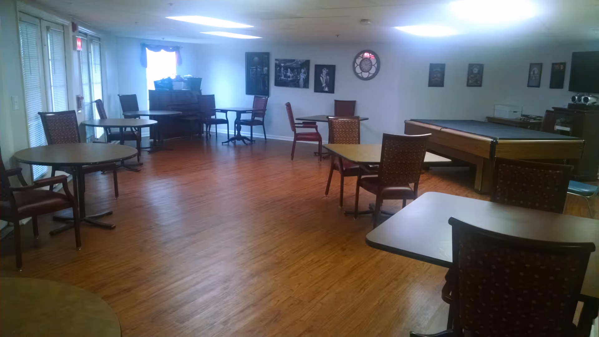A common room with several round and square tables surrounded by chairs, a piano against the far wall, a pool table, and framed pictures and a clock on the walls. The room has wooden flooring and is lit by ceiling lights.