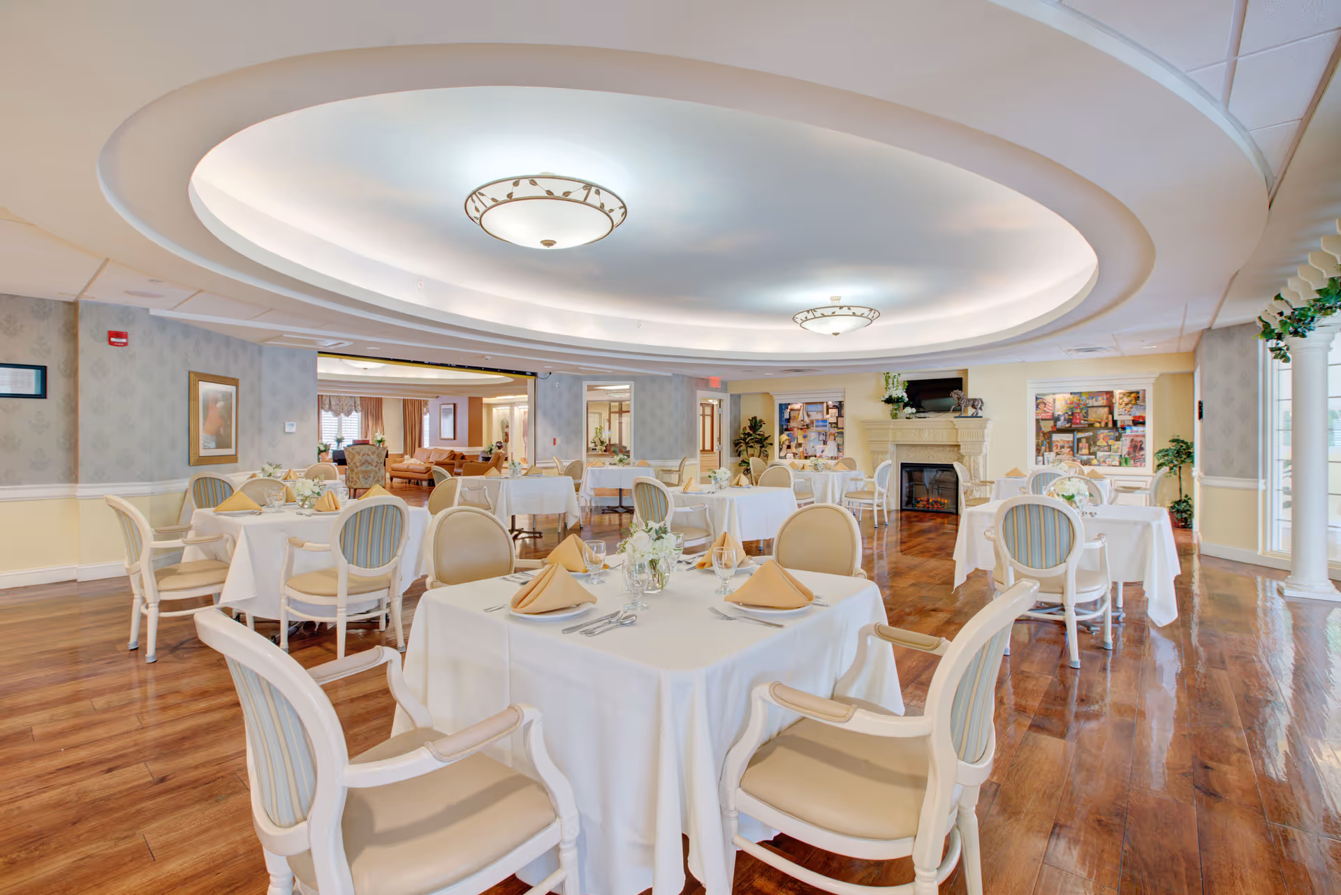 Bright, elegant dining room with white-tablecloth tables set with napkins and chairs beneath a large recessed ceiling feature and a fireplace in the background.
