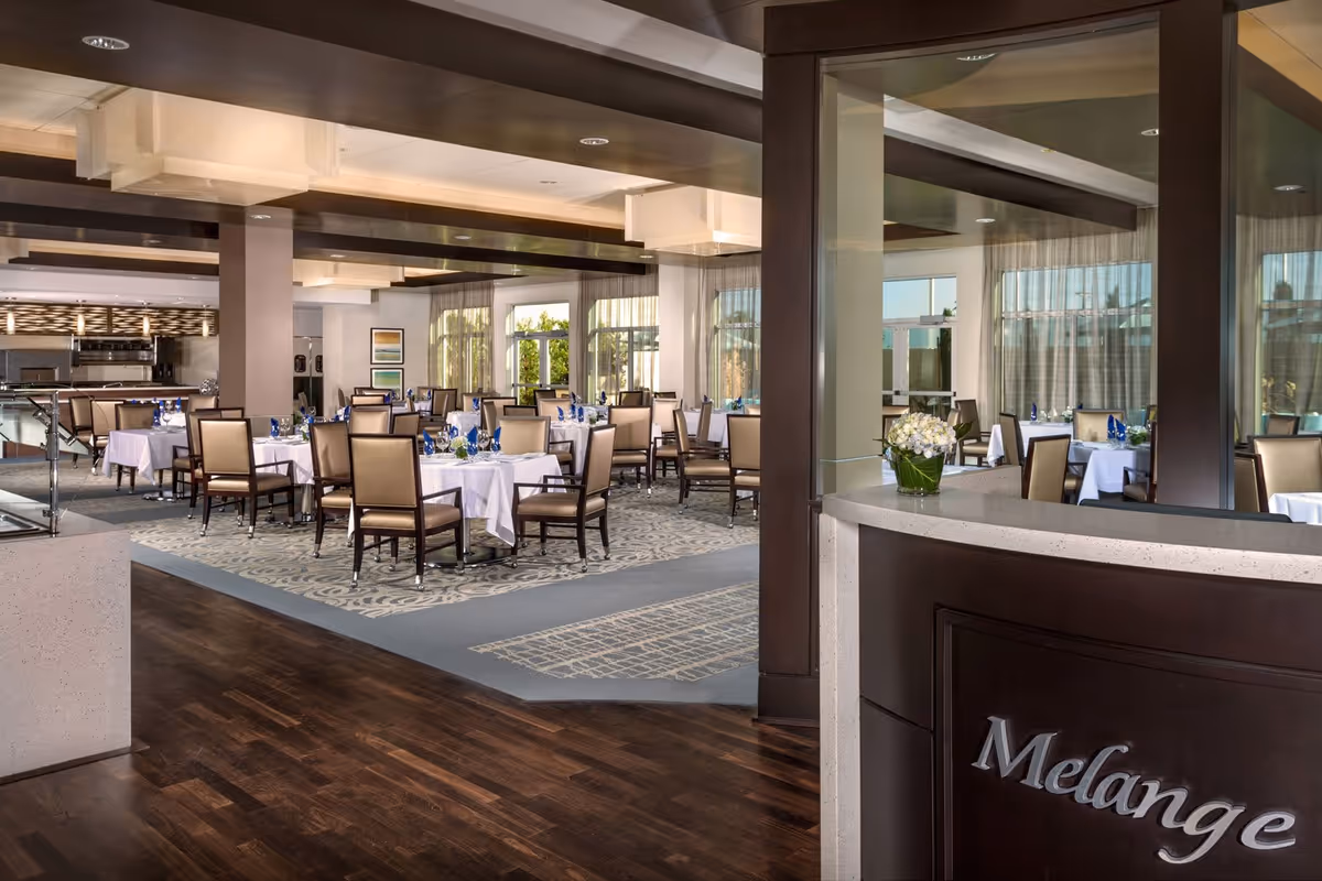 A spacious and elegant dining room with multiple tables covered in white tablecloths, each set with blue napkins and glassware. The room features large windows with sheer curtains allowing natural light to fill the space. The floor is a combination of dark wood and patterned carpet. In the foreground, there is a reception desk with the word 'Melange' displayed on the front and a vase of white flowers on top.