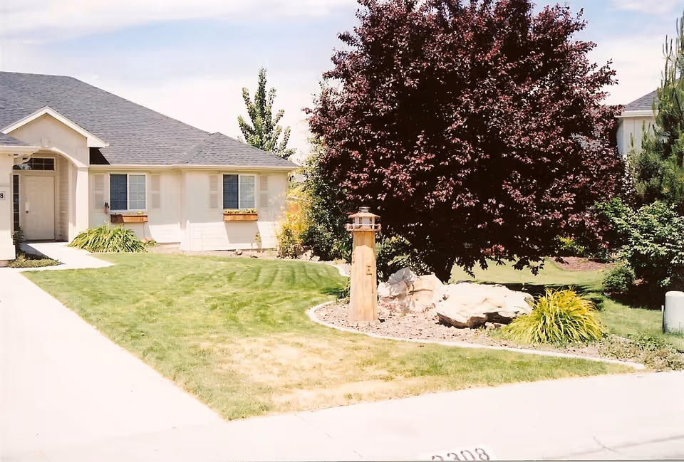 Exterior view of a single-story building with a gray roof and beige walls, surrounded by a well-maintained lawn and landscaped garden featuring a large tree, rocks, and shrubs. A concrete walkway leads to the entrance of the building.