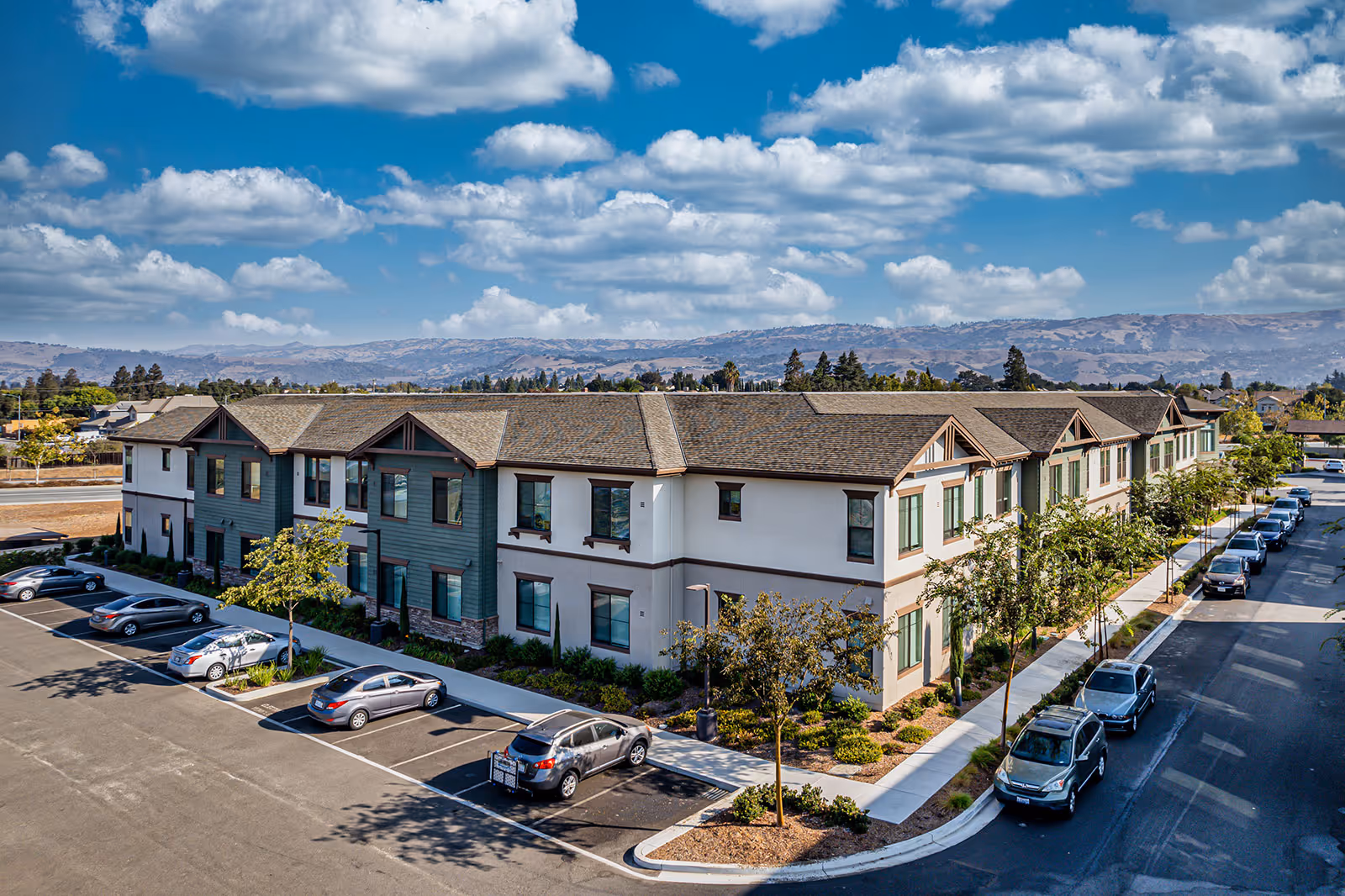 Two-story residential senior living building with parked cars, landscaped sidewalks, and distant hills under a partly cloudy sky.