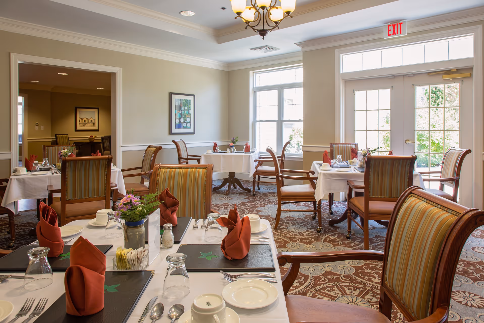 A bright dining room with several tables set for a meal, featuring white tablecloths, folded red napkins, plates, glasses, and silverware. The room has large windows and glass doors letting in natural light, striped upholstered chairs, a patterned carpet, and a chandelier hanging from the ceiling.