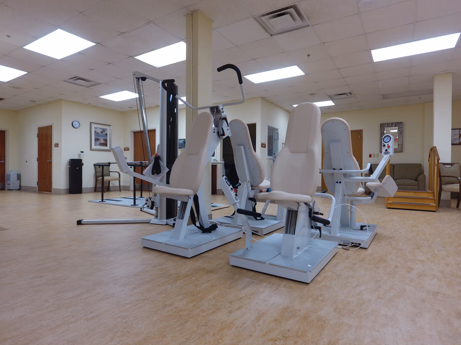 A spacious room with exercise equipment designed for seated workouts, including machines with padded seats and handles. The room has a light-colored wood floor, beige walls, fluorescent ceiling lights, and several doors. There are chairs and a small staircase with handrails in the background.