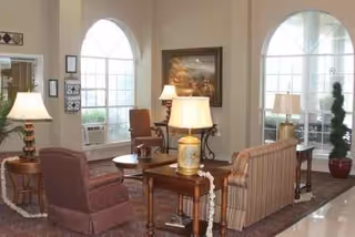 Cozy senior living lounge with cushioned seating, tables, lamps, and arched windows.