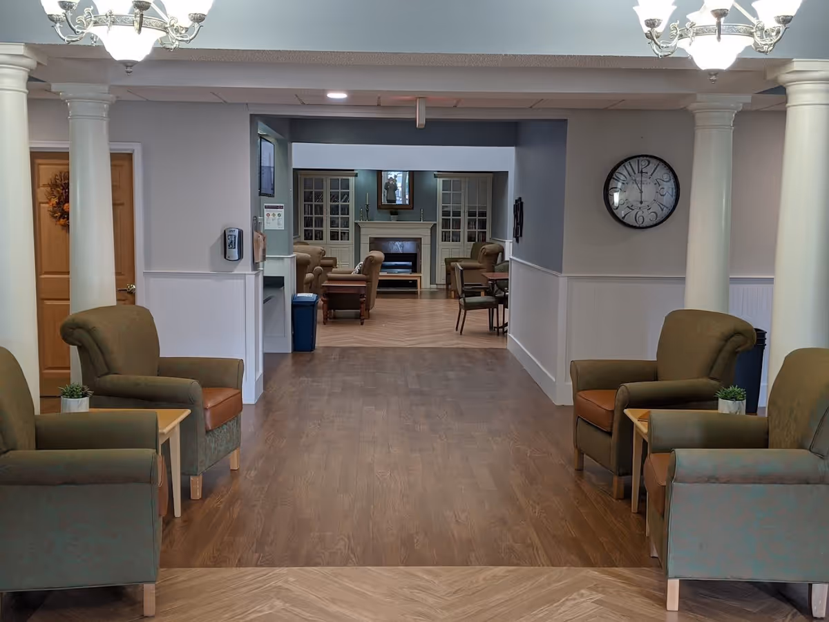 Interior view of a senior living facility lounge area with green armchairs and small tables arranged along a wooden floor hallway. The hallway leads to a cozy sitting area with more armchairs, a fireplace, and built-in cabinets. The walls are painted light gray with white wainscoting, and there are two white columns and chandeliers visible in the foreground. A large wall clock is mounted on the right wall.