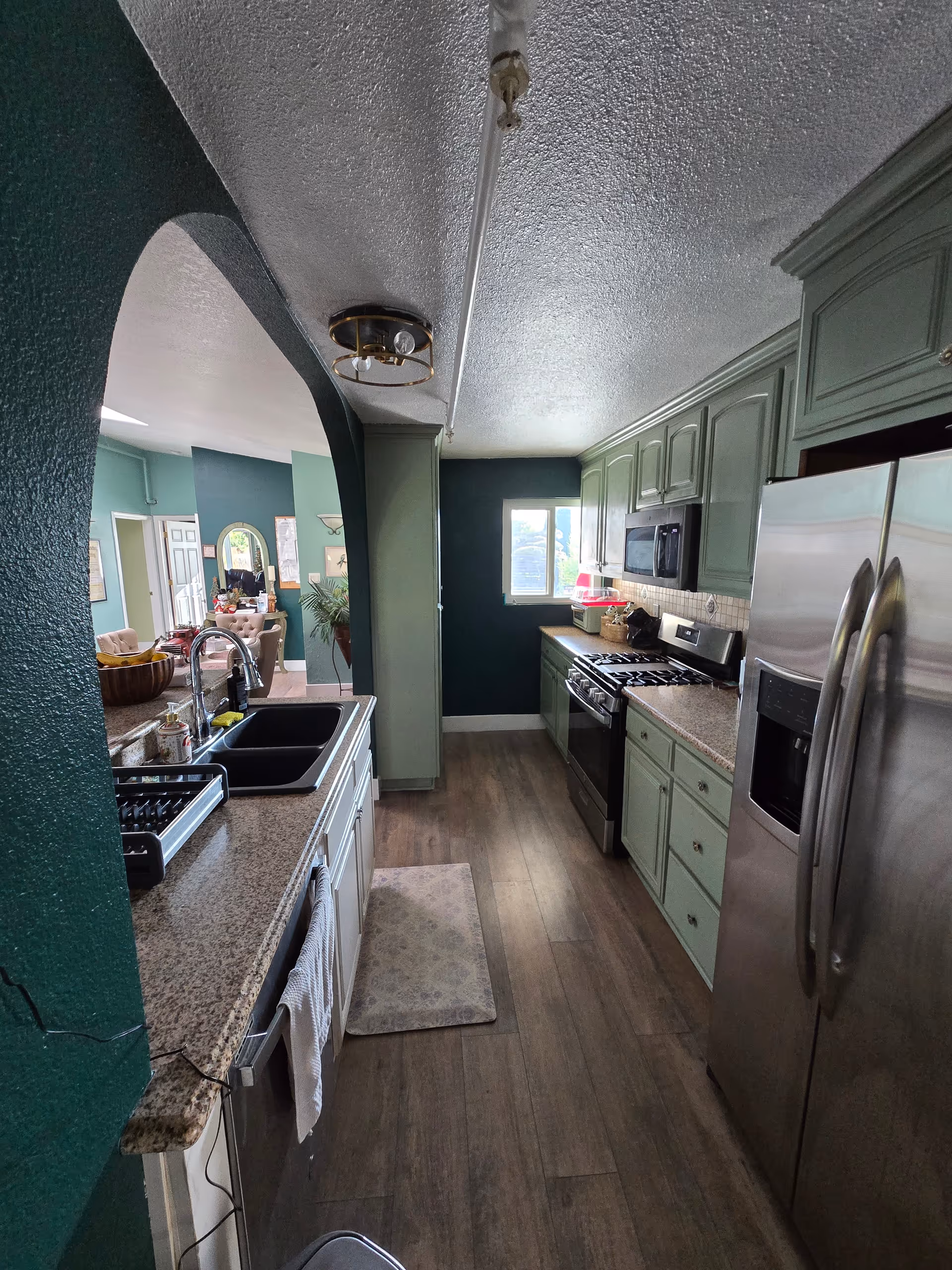 A narrow kitchen with green cabinets, granite countertops, a double sink, stainless steel refrigerator, stove, and microwave. The kitchen has wood flooring and a small window at the far end. The kitchen opens into a living area with green walls and visible furniture.