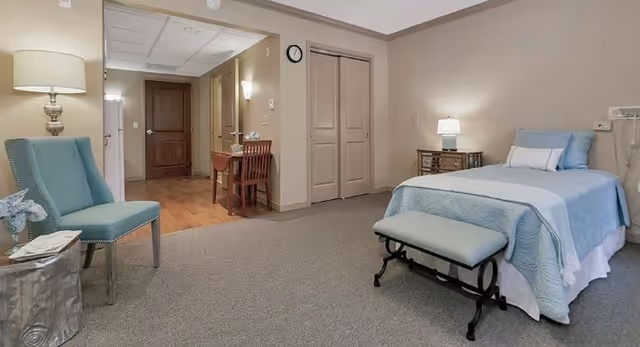 Private senior-living bedroom with a single bed, bench, armchair, side tables, a desk, and an open hallway to the entry.