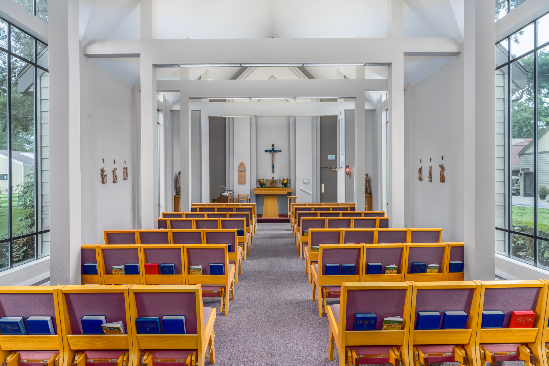 Interior view of a small chapel with rows of wooden pews with purple cushions and hymnals. The chapel has large windows on both sides, white walls, and a crucifix mounted on the front wall above a small altar with flowers and religious items.